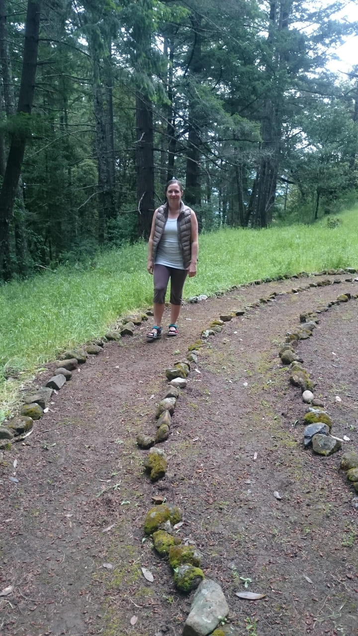 Johanna stands on a forest path lined with moss-covered stones, dense trees rising behind her.