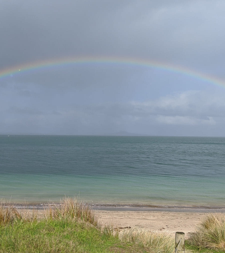 A wide double rainbow arches across a calm ocean and overcast sky, with a small boat near the left shore and a quiet sandy beach in the foreground.