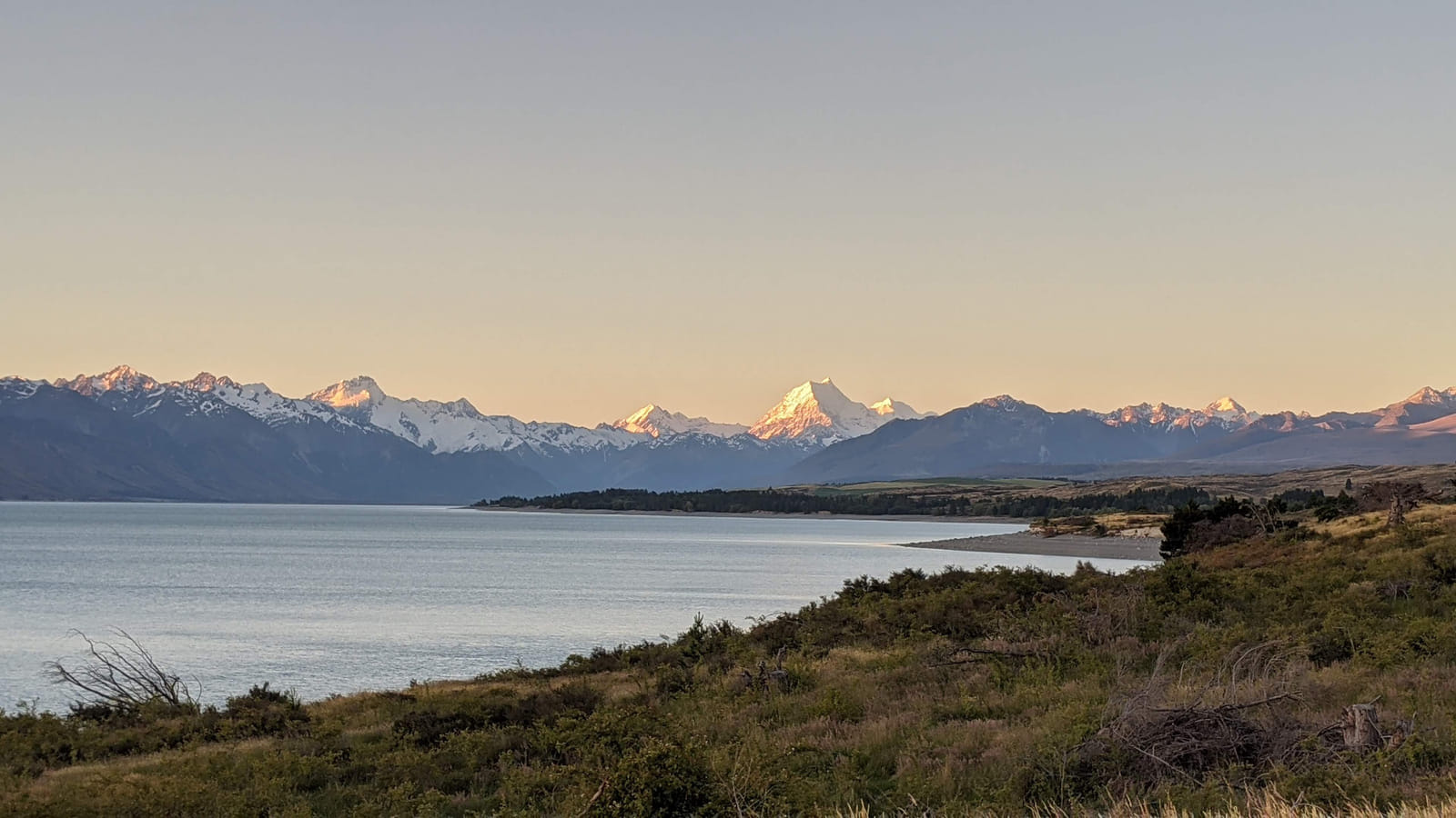 An alpine lake at dawn with golden light on snow-capped mountains across the water; soft warm lighting and muted vegetation along the shoreline.
