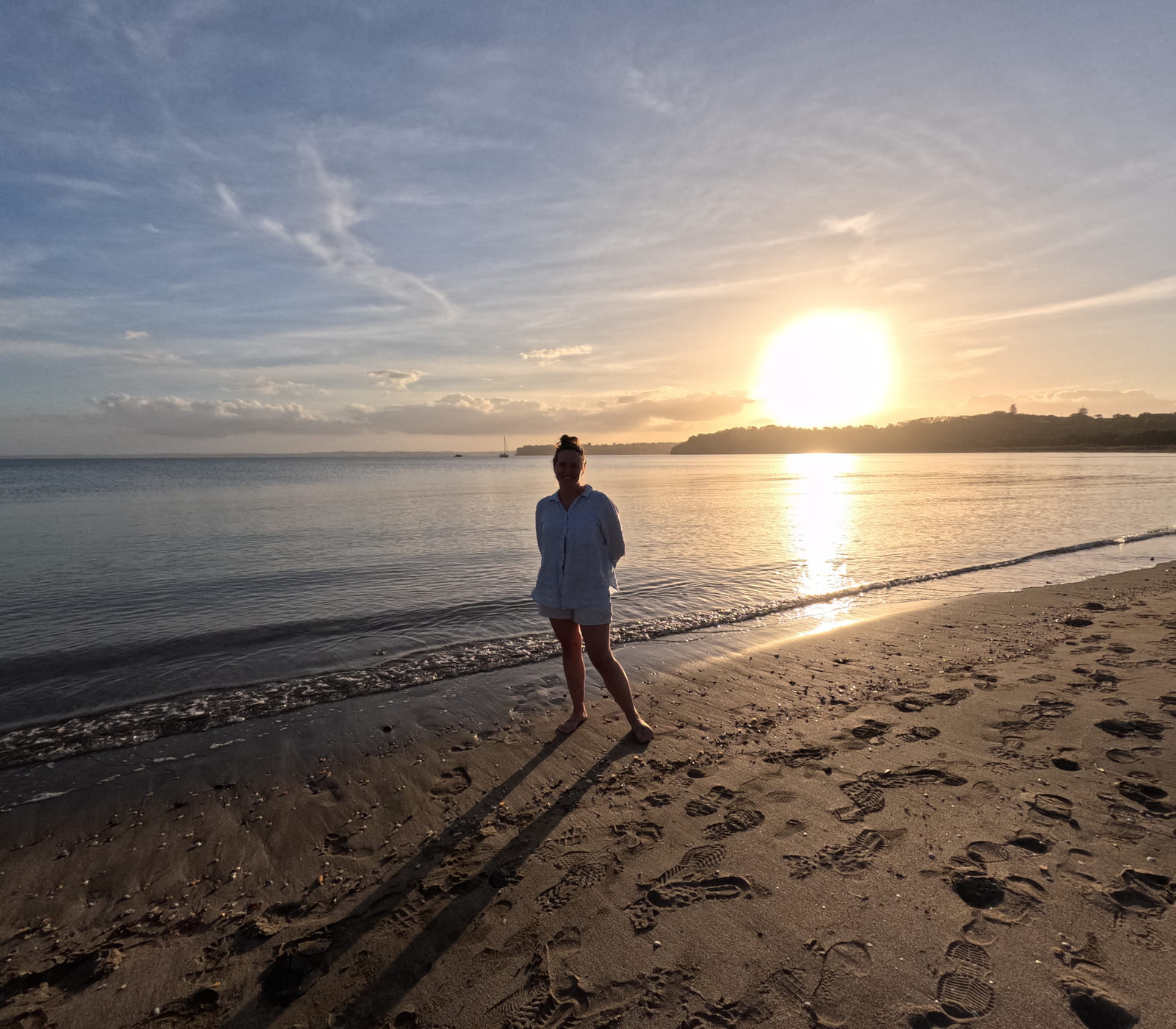 Johanna walks along a sandy beach at sunset, seen from behind as a silhouette against the brilliant setting sun and golden reflections on wet sand.